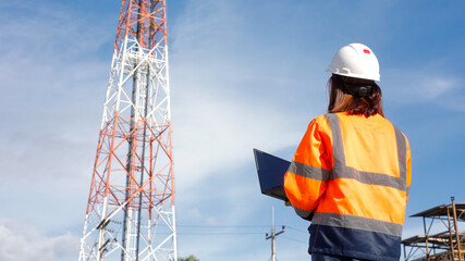 A worker in safety gear stands with a laptop, looking up at a tall telecommunications tower. The bright sky adds to the scene, showcasing a busy work environment