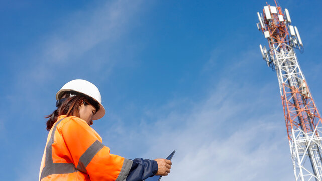 A female technician in a safety vest and hard hat inspects her tablet while standing near a tall telecommunications tower under a clear blue sky