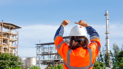 A construction worker in a bright orange safety jacket and helmet stands with arms raised at an industrial site. Tall structures and a clear blue sky are visible, indicating activity