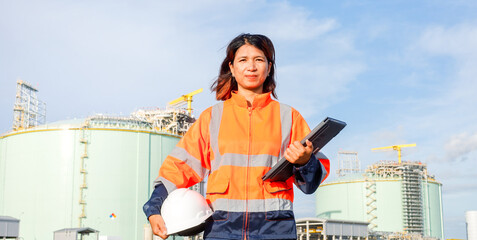 A confident woman stands at a construction site wearing an orange safety jacket and helmet. She holds a clipboard while overseeing large storage tanks and equipment under a clear blue sky © Happy Photo