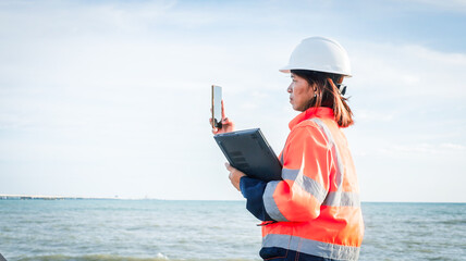 A worker wearing a hard hat and reflective clothing stands by the ocean. She is taking photos of the construction site, assessing the area under bright, clear skies © Happy Photo