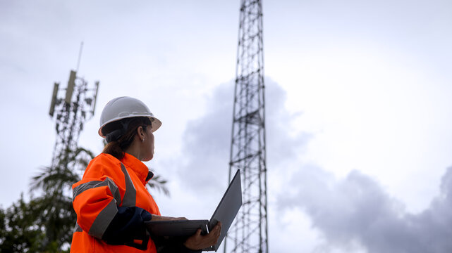 A worker in bright orange safety clothes and a hard hat stands with a laptop in hand, looking up at a tall telecommunications tower against a cloudy sky