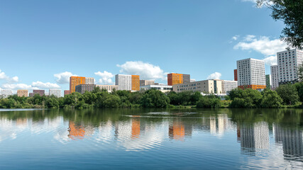 Modern residential buildings with colorful facades reflected in a calm river surrounded by green trees under a bright blue sky.