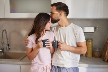 In a bright kitchen, a couple shares a warm moment while holding mugs, smiling, and exchanging...
