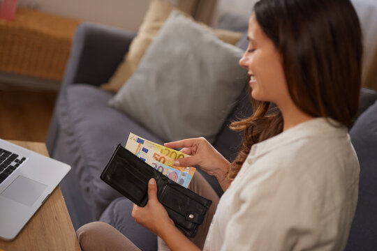 A woman is sitting comfortably on a couch, sorting euro banknotes from her wallet. A laptop is open on the table in front of her, creating a relaxed atmosphere.