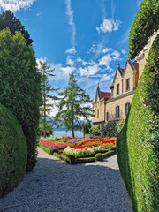 Oberhofen Castle garden on Lake Thun with colorful flower beds, a stone path leading to the historic castle, and scenic lake view in the background on a sunny day.