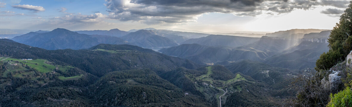 Panoramic view taken from Mirador del Far, near the medieval village of Rupit. A landscape of mountains, hills, and valleys covered in forests, with green areas illuminated by the sunset light. Spain