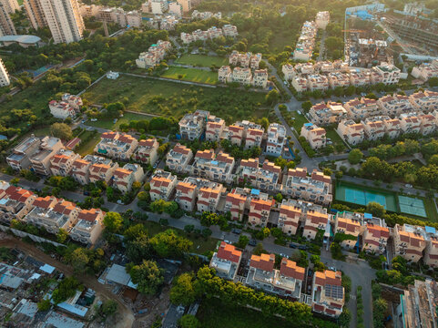 Aerial view of symmetrical rows of terracotta-roofed houses nestle amidst verdant trees, creating a tapestry of warm hues against the urban sprawl, Gurugram, Haryana, India.
