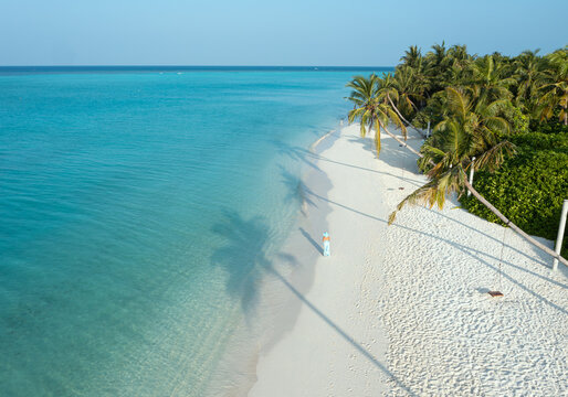 Aerial view of a lone figure strolls along a pristine white beach, where turquoise waters meet long palm tree shadows, Thoddoo, Alif Alif Atoll, Maldives.