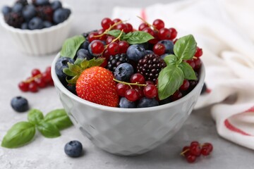Different ripe berries and basil leaves in bowl on light grey textured table, closeup