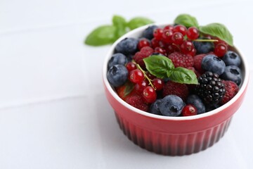 Different ripe berries and basil leaves in bowl on white tiled table, closeup. Space for text