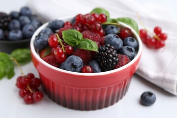 Different ripe berries and basil leaves in bowl on white table, closeup