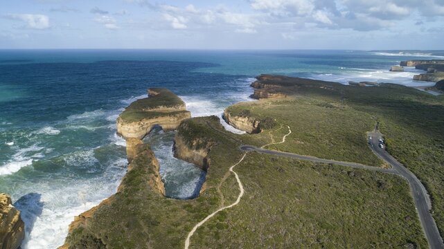 Aerial view of rugged cliffs meet the turbulent ocean, with pathways winding through the green landscape, Port Campbell, Victoria, Australia.