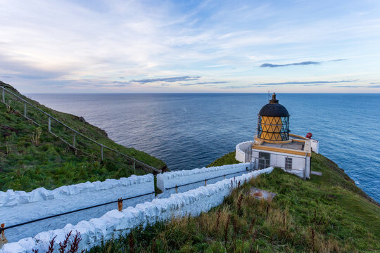 View of the lighthouse standing proudly against the vast, undulating blue sea under the cloudy sky, with a white stone path leading to it, St Abbs Head, Scotland.