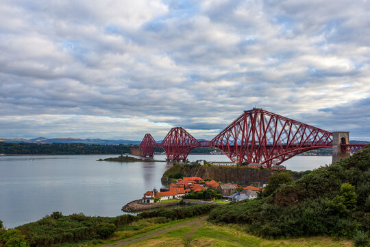 View of the iconic, crimson Forth Bridge stretches across the tranquil Firth of Forth under a textured sky, reflecting in the still water, Dalmeny, Scotland.