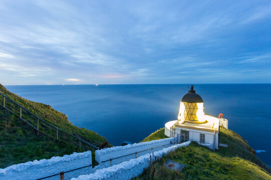 View of a radiant lighthouse perched atop rugged cliffs, its light cutting through the twilight over the serene, dark blue sea, St Abbs Head, Scotland.