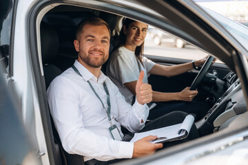 Gesture of thumb up, smiling. Woman is with instructor in car, driving school concept
