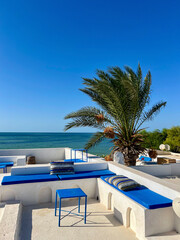 Enjoy the stunning scenery from a cozy rooftop in Djerba, Tunisia. The whitewashed terrace features blue seating, offering a perfect view of the sea and nearby palm trees.