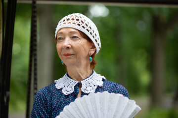 Older woman in vintage attire stands with a fan amidst lush greenery in a tranquil outdoor setting