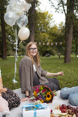 Smiling pregnant woman sitting on picnic blanket in park with balloons, flowers, and gifts during baby shower celebration. Concept of maternity lifestyle, family happiness, and outdoor party. Symbol