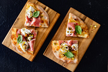 Slices of Pinsa Romana with mortadella, burrata cheese and pistachio on wooden cutting boards on black background