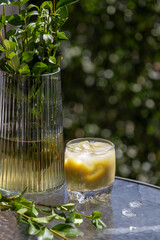 An iced matcha latte in a glass, accompanied by fresh green leaves in a vase, sits on an outdoor table bathed in bright sunlight. This refreshing drink is ideal for a warm summer day.