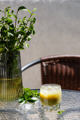 A vibrant iced matcha drink sits on a glass table next to a vase of green foliage, with scattered ice cubes and fresh leaves. This refreshing beverage is perfect for a sunny outdoor setting.
