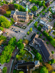 Aerial view of a European town with a tall green spire gothic church at its center, surrounded by red roofed houses, winding streets, trees, and fields blending into rural outskirts.