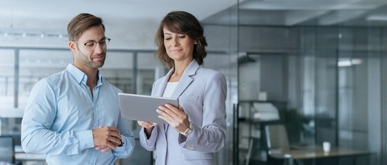 Mature mentor woman and European young man discussing work on tablet in office. Two diverse...