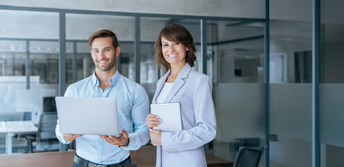 Obraz premium Team portrait at camera of partners mature Latin businesswoman, European business man discussing project on laptop in office. Two professional people mentor and intern working. Banner, copy space