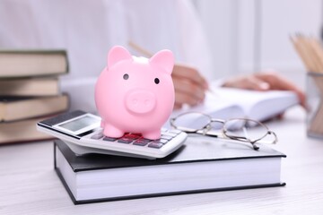 Piggy bank with calculator, book on white wooden table and woman working indoors, selective focus