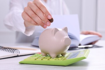 Woman putting coin into piggy bank at white wooden table, closeup