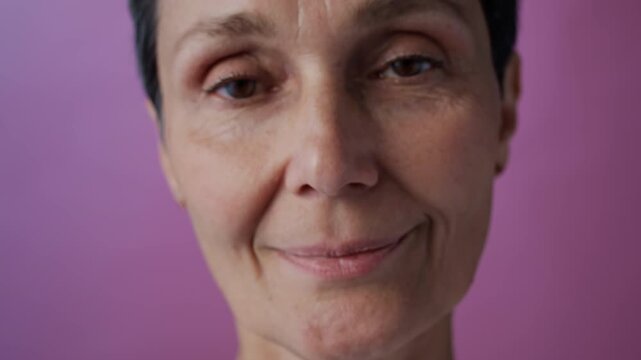 Rack focus of mature Caucasian woman, breast cancer survivor, holding pink ribbon looking at camera with smile on pink background, female solidarity concept