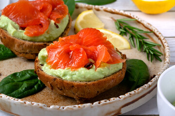 Bruschetta with smoked trout fish and avocado cream on bread served on the ceramic plate with lemon and herbs