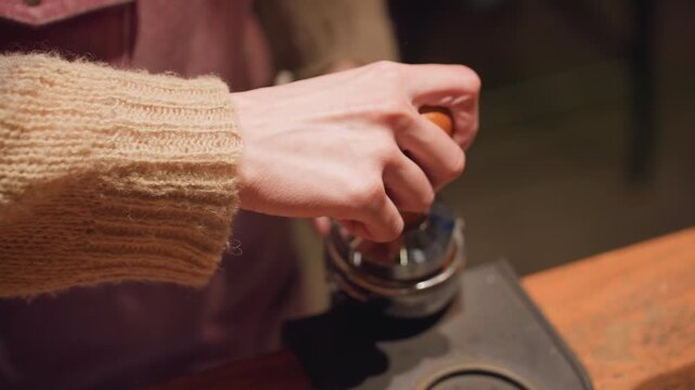 Close up of bartender using tamper to press coffee grounds into portafilter with focused hand movement over bar counter, warm lighting and soft textures