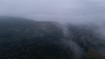 Fototapeta premium Aerial of Village in rain cloud cover tropical green mountain. Rainy season. Misty cover green forest. beautiful green village