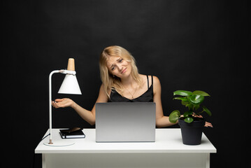 Woman sitting at desk with laptop shrugging shoulders against black background