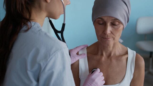 Tilt down shot of young female therapist auscultating lungs of mature woman with cancer using stethoscope in clinic