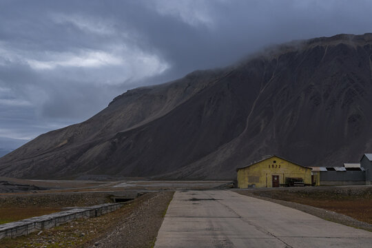 View of a stark, abandoned building stands under a brooding sky, overshadowed by a colossal mountain, a path leading towards its silent facade, Pyramiden, Svalbard and Jan Mayen.