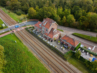 Fototapeta premium Aerial view of a red roofed cafe beside railway tracks and a road crossing with safety barriers, set in a green semi rural landscape of trees and vegetation.
