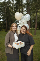 Smiling pregnant woman standing with friend in park holding box of cupcakes and balloons during baby shower celebration. Concept of maternity lifestyle, friendship, and family happiness. Symbol of