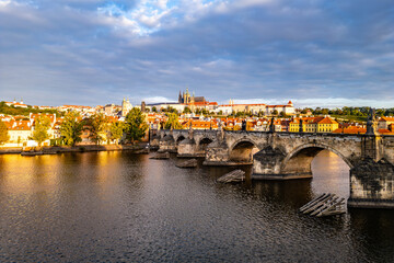 Early morning light casts a glow on Prague Castle and Charles Bridge, reflecting beauty over the Vltava River. The old town awakens as clouds drift above the historic skyline.