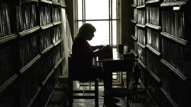 A woman sits in a semi-dark room of a book depository and sorts through catalog cards. Shelves with books on both sides, working with library archives, reading alone
