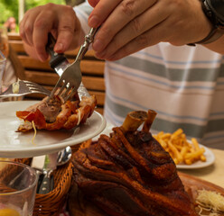 A close-up of hands carving roasted pork knuckle onto a plate. Golden crispy skin, tender meat, and side dishes create an authentic dining experience. The rustic presentation reflects traditional cuis