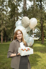 Smiling pregnant woman standing outdoors with festive balloons and a box of cupcakes during baby shower celebration. Concept of maternity lifestyle, family happiness, and pregnancy event. Symbol of