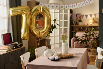 Birthday party scene showing large golden 70 balloon and happy birthday banner in decorated living room, table set with teapot, cookies, pastries, vase with flowers, no people visible
