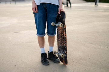 A young person stands in a park during a warm summer evening, holding a skateboard and looking ready to enjoy some skating fun with friends nearby. © Mihail