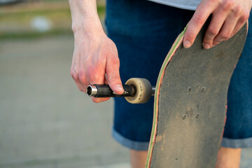 A person is working on their skateboard, using a fix tool to adjust a wheel while standing on a paved area, enjoying the outdoor atmosphere.