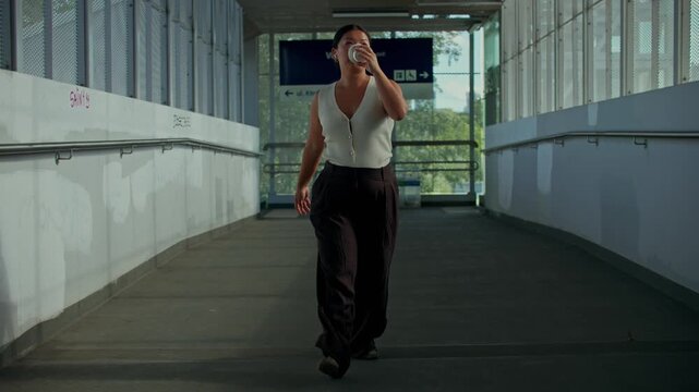 A woman is talking on a mobile phone through earbuds and drinking coffee walking along the train station.