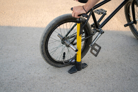 Rider uses a hand pump to inflate the tire of a BMX bicycle on a gravel surface with sunlight providing a bright atmosphere.